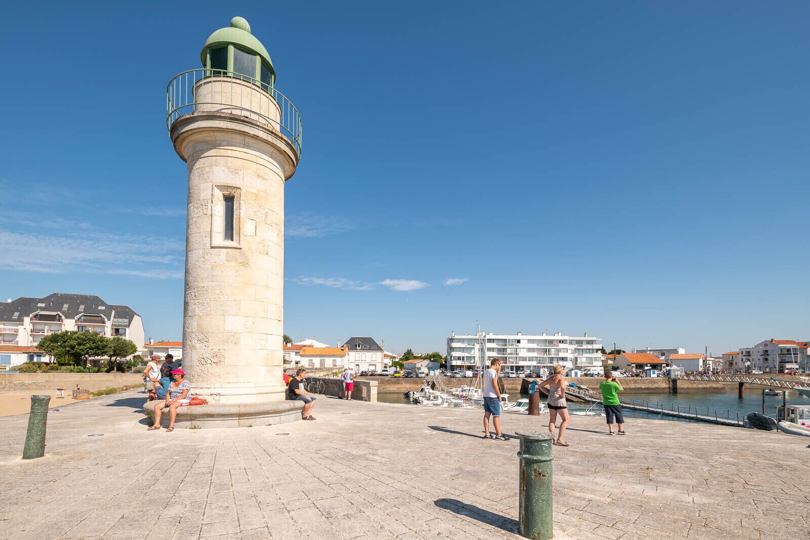 A lighthouse with a circular base and a dome on top, situated on a paved area with people around.