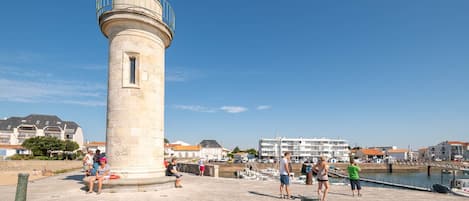 A lighthouse with a circular base and a dome on top, situated on a paved area with people around.