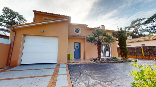 Une maison à deux étages avec un garage, une terrasse couverte et un petit jardin agrémenté d’un palmier.