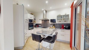 A modern kitchen with white cabinets, a stainless steel range hood, a built-in oven, a coffee maker, and a dining area with two black chairs.