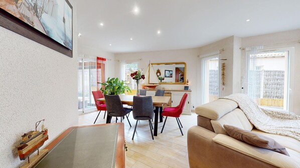A modern living room with a sofa, dining table, and chairs. There is a large window providing natural light and a view of the outside.