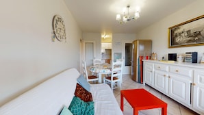 A cozy living room with a sofa, a dining table with chairs, a kitchen area with cabinets, and a red bench. - Nautica - 1-Bedroom Apartment Near the Beach & Parking (Saint-Hilaire-de-Riez)