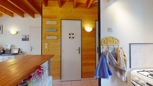 A room with wooden ceiling beams, a white door with signs for 'Pornichet', 'La Baulde', and 'Guérande', a life preserver on the door, a towel rack with towels, a wooden table with wine glasses, and a white stove. - Escapade Turballaise - House 400m from the Port (La Turballe)