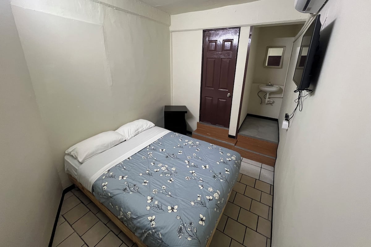 Une chambre à coucher avec un lit, une porte, une petite table, un téléviseur et une salle de bain visible à travers une porte ouverte.