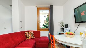 Een knus woongedeelte met een rode bank, een eettafel met een schaal fruit en een aan de muur bevestigde televisie.