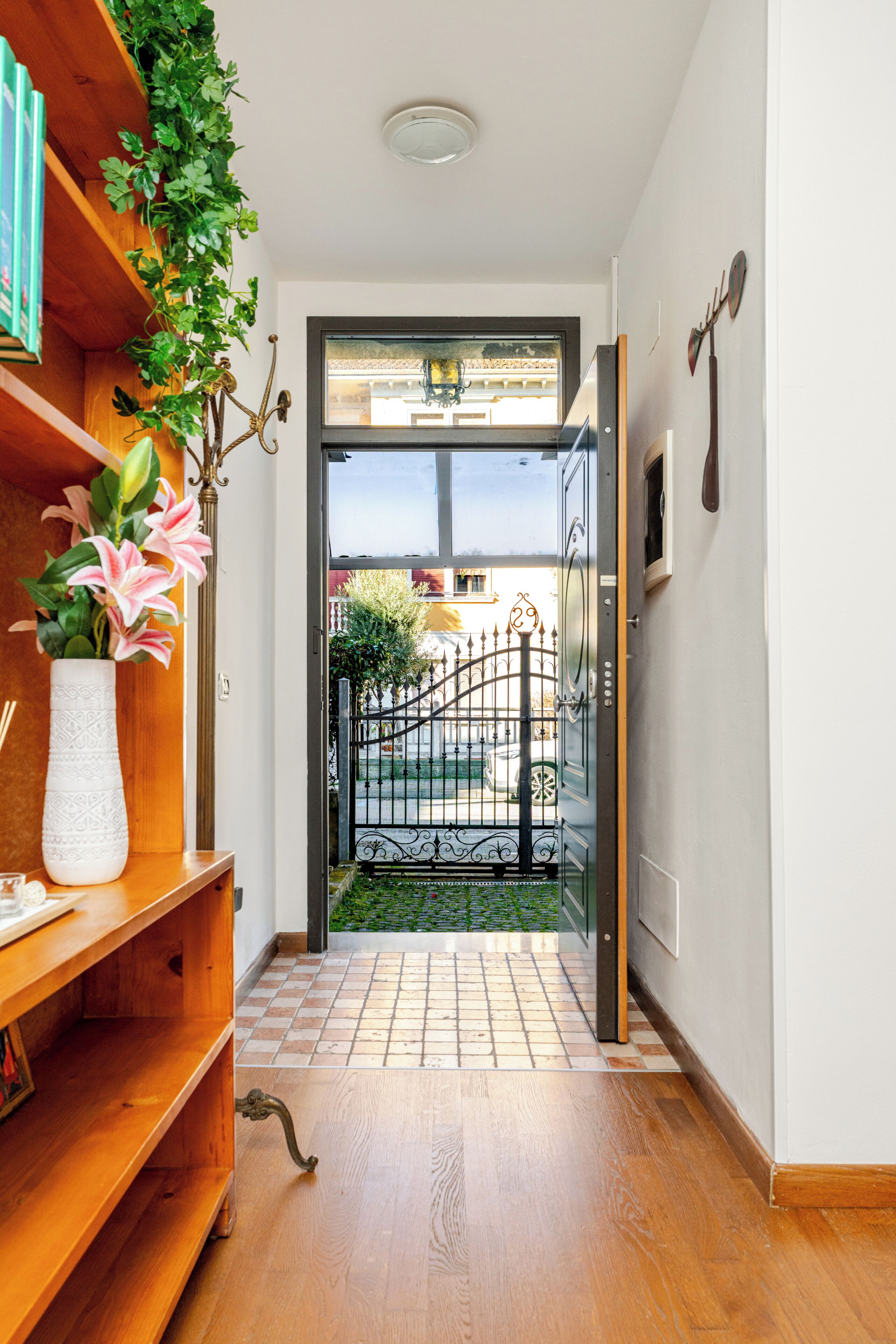 A hallway with a wooden floor, a potted plant, a metal gate, and a door leading outside.