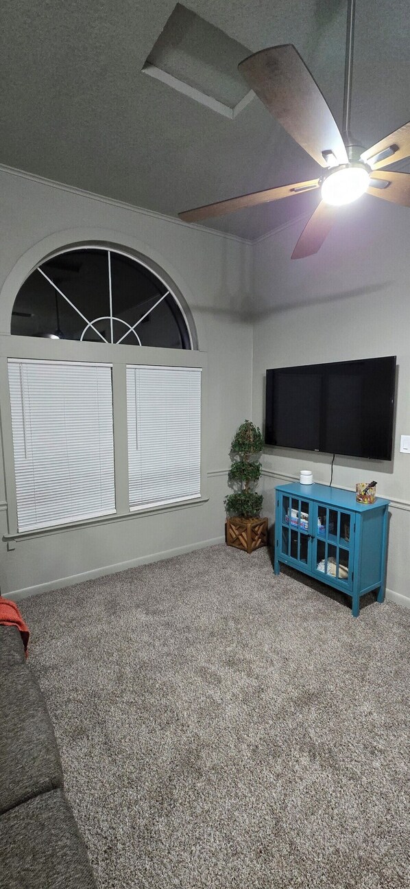A room with a ceiling fan, a large window with blinds, a flat-screen TV, a blue cabinet, and a small plant.