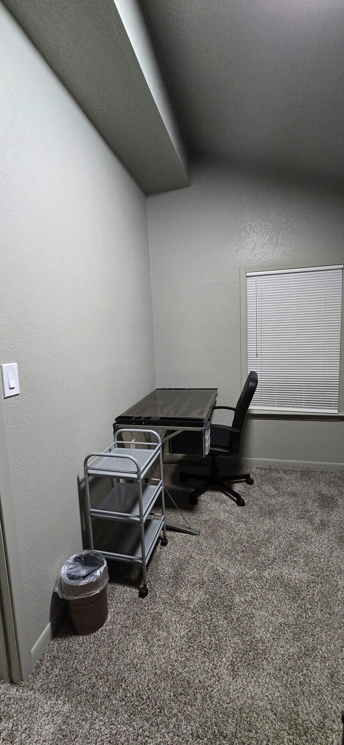 A room with a carpeted floor, a table, a chair, a trash can, and a window with blinds.
