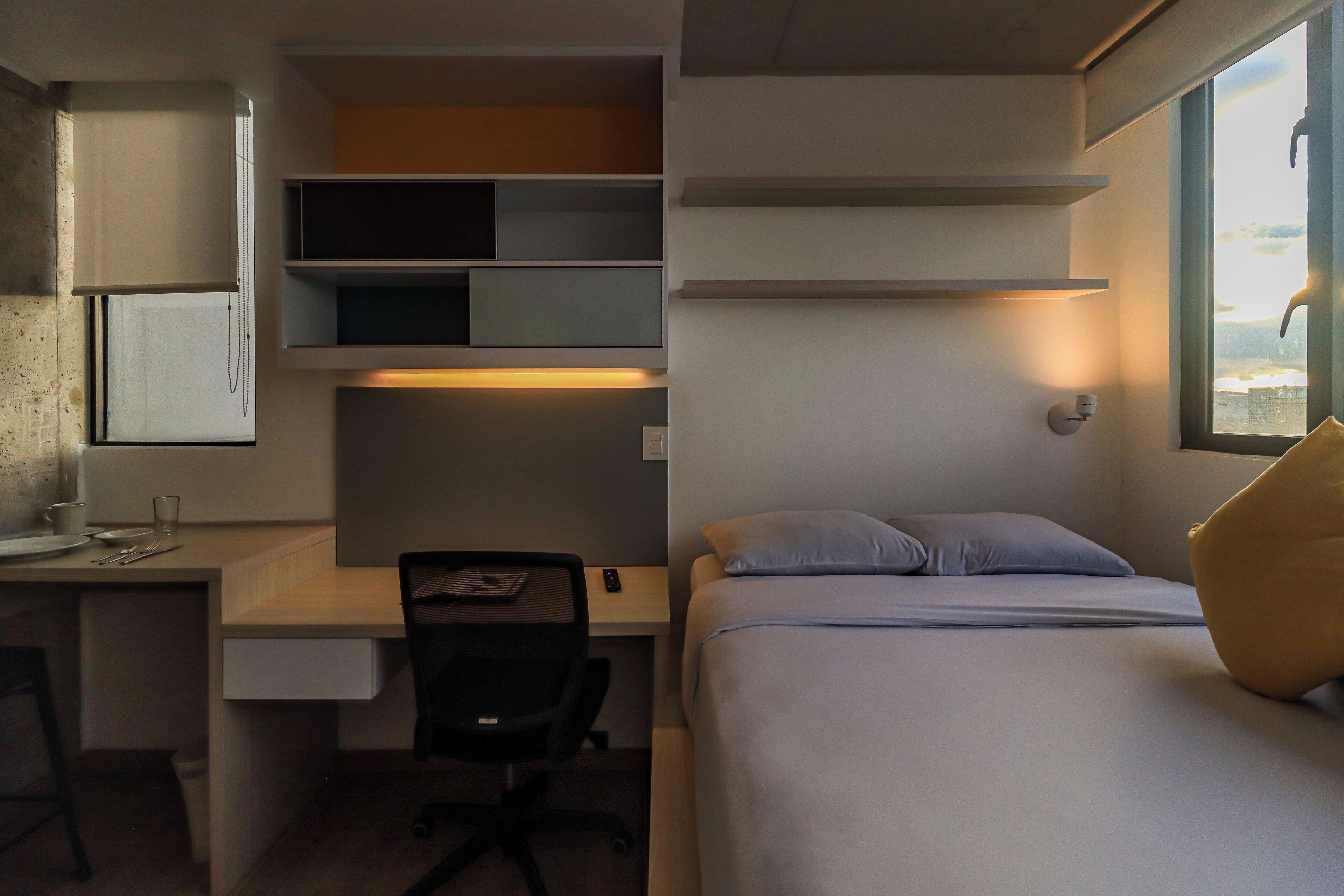 A compact hotel room with a bed, a desk with a chair, a small kitchenette, and a window with a view.
