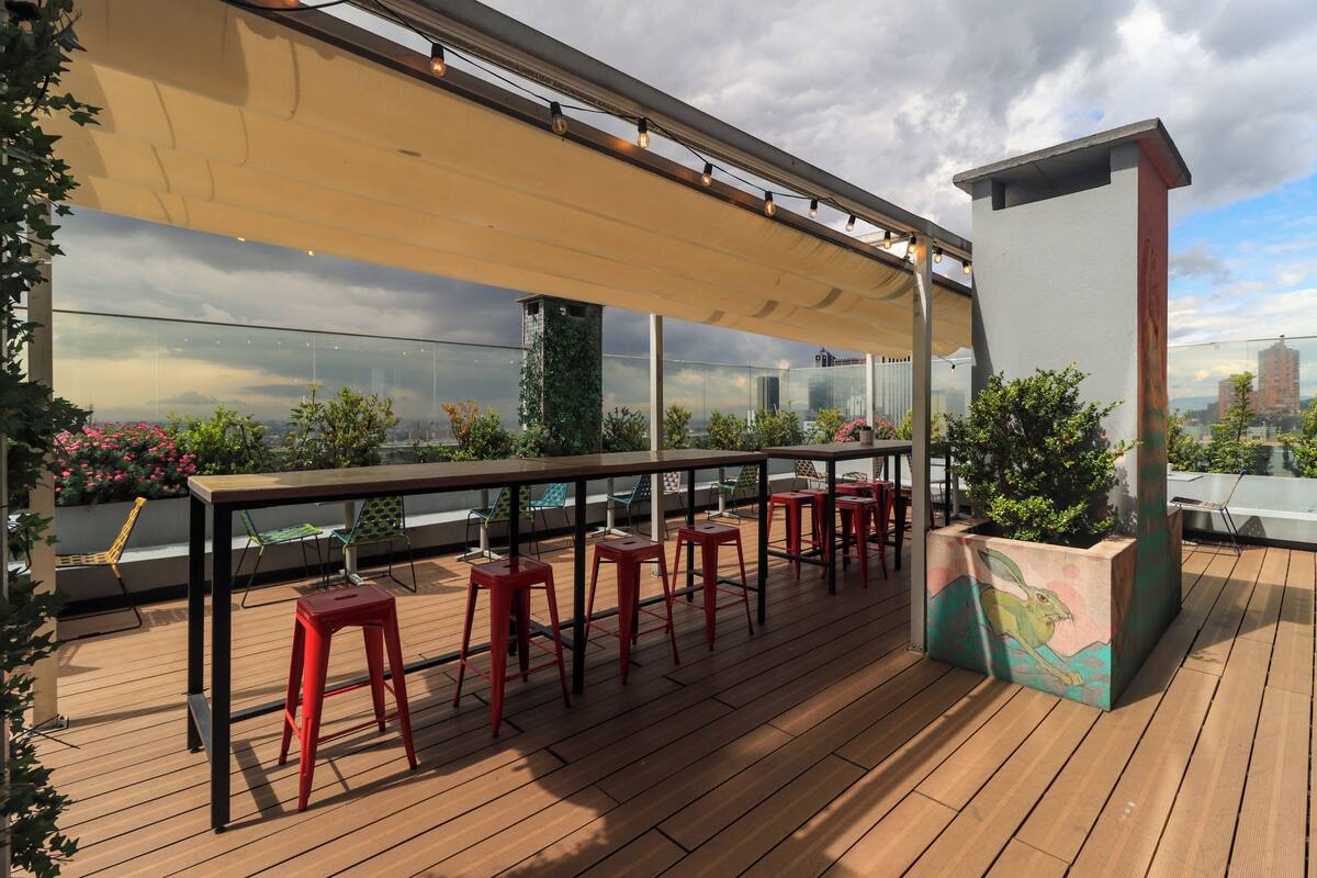 Terraza en la azotea con cubierta de madera, taburetes de bar rojos, una mesa y un ventanal con vistas a la ciudad.