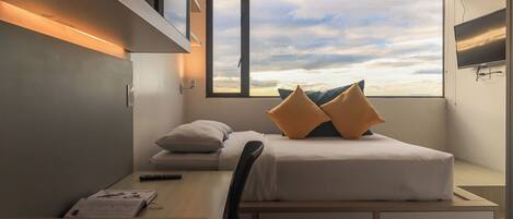 A compact, modern room with a bed, a desk, and a window offering a view of the sky.