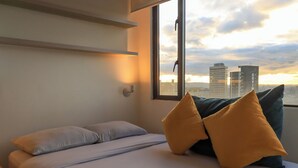 Quarto com cama, almofadas, mesa de cabeceira, janela e vista para a paisagem urbana.