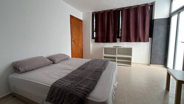 A bedroom with a bed, a wooden door, a window with curtains, a white storage unit, and a small table.