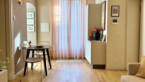 A living room with a dining area, a sofa, a coffee table with books, a kitchen area, and a large window with curtains. - Residenza Della Bollente II - Stylish cosy apartment in the heart of the town. (Acqui Terme)