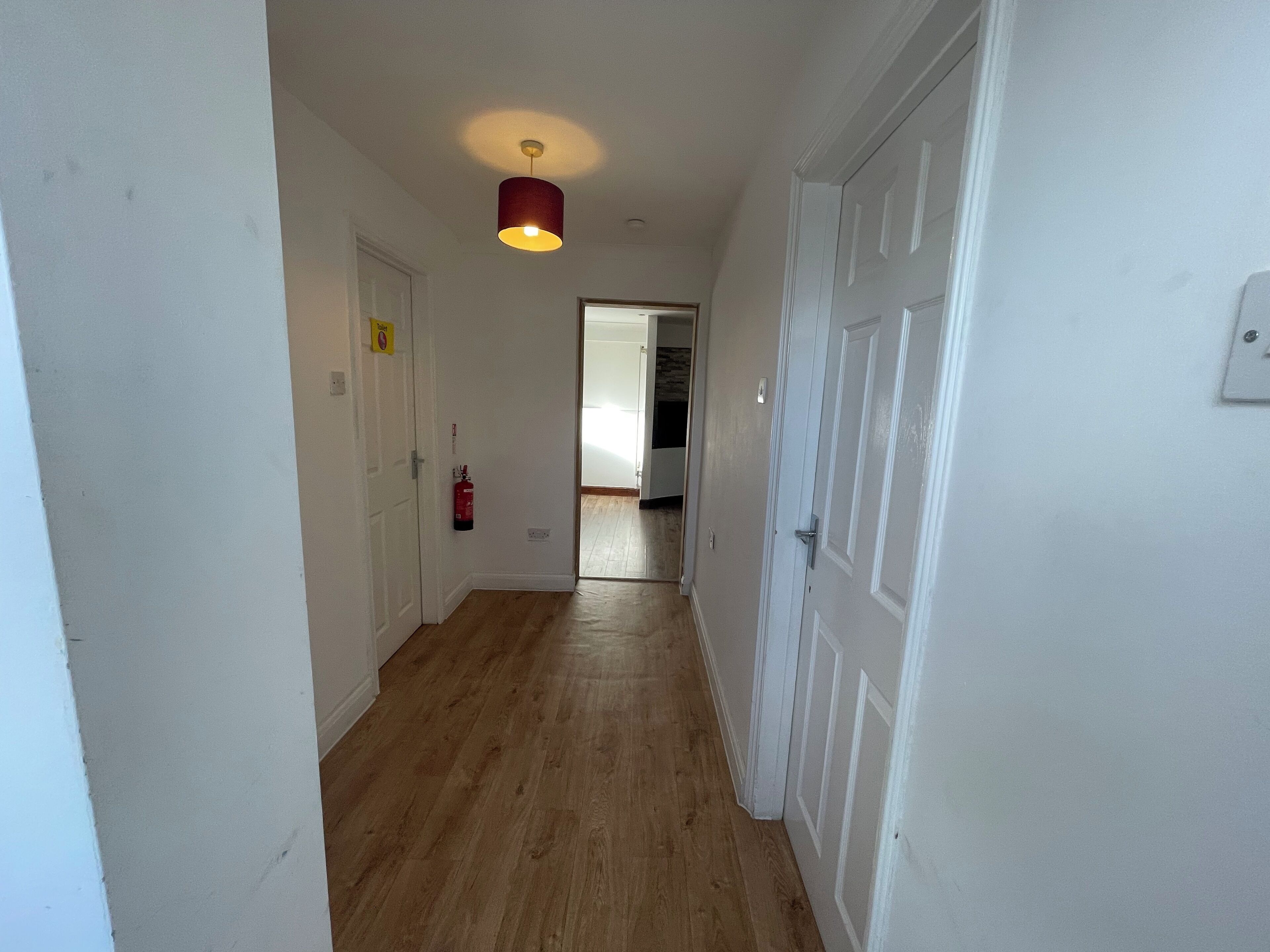 A hallway with wooden flooring, white walls, and a red pendant light. There is a fire extinguisher mounted on the wall and a door on the right.