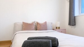 A neatly made bed with a white comforter and two rolled towels, a bedside table with a lamp, and a window with a curtain. - Peaceful 4-bedroom house in quaint stevenage perfect for families & contractors (Stevenage)