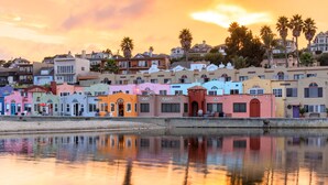 Una propiedad frente al agua con edificios de colores, una superficie acuática tranquila y reflectante, y palmeras al fondo.