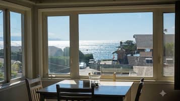 A dining area with a table and chairs, large windows with a view of the ocean and buildings.