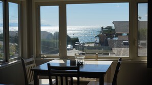 Comedor con mesa y sillas, ventanas amplias con vista al océano y edificios.