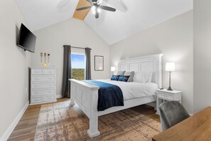 Ein Schlafzimmer mit Bett, Nachttisch, Schreibtisch, Stuhl, Fernseher, Deckenventilator und einem Fenster mit Vorhängen.
