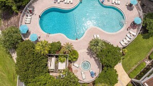 Une vue aérienne d’une piscine au design original, avec des formes ondulées, entourée de chaises longues et de parasols.