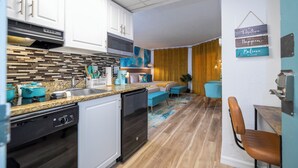 Une cuisine moderne avec des meubles blancs, un lave-vaisselle noir et un plan de travail en granit. On y trouve également un coin repas avec une chaise bleue et une table.