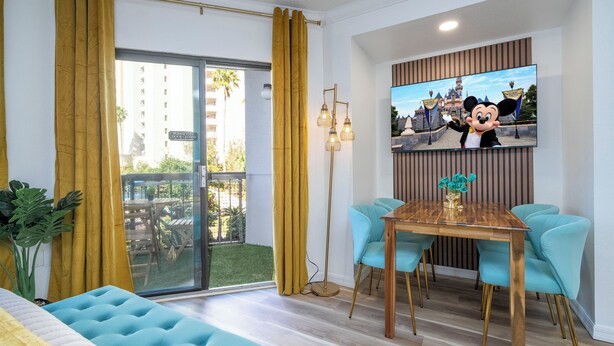 Une chambre d’h ôtel avec un balcon, un coin repas avec une table en bois et des chaises bleues, un grand téléviseur fixé au mur et une plante sur la gauche.