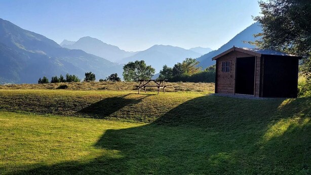 Ein rustikales Steinhaus mit einer Holzkonstruktion in der Nähe, vor einer Bergkulisse und einer Wiese.