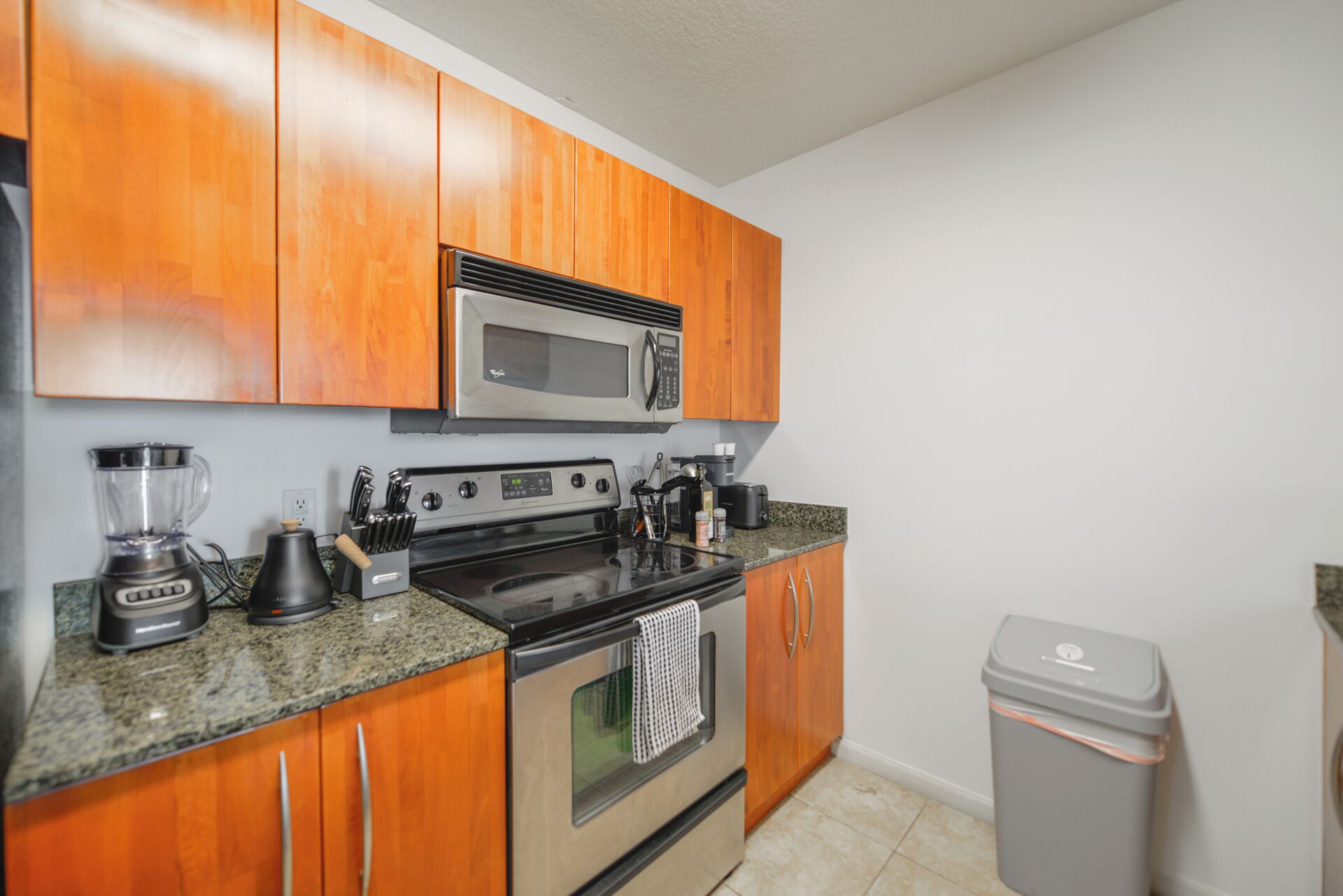 A kitchen with wooden cabinets, a microwave, a stove, a blender, and a trash can.
