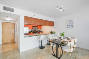 Eine moderne Küche mit Holzschränken, einem Edelstahlkühlschrank und einer Mikrowelle. Es gibt einen Essbereich mit Tisch und Stühlen sowie einen kleinen Topf auf dem Tisch.