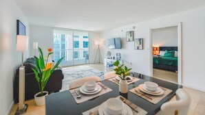 A modern living room with a dining area, a sofa, a TV, and a balcony with a city view. - Brickell Bay 3318 (Miami)