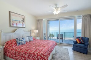 A bedroom with a large bed, a ceiling fan, a view of the ocean through a sliding glass door, and a blue armchair. - Princess Condos - All Units Are Gulf Front & Direct Beach Access in PCB! (Panama City Beach)