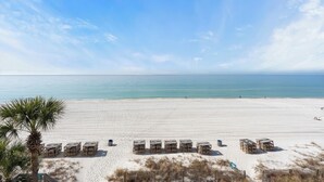 En strand med hvidt sand, en klar blå himmel og et roligt hav. Der er trækonstruktioner i forgrunden, muligvis til strandaktiviteter.