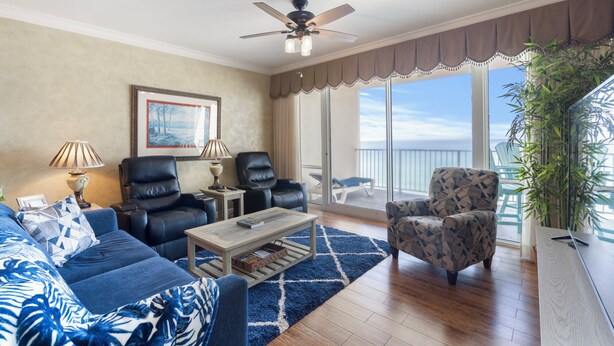 A living room with a sofa, two chairs, a coffee table, and a ceiling fan. There is a large window with a view of the ocean. - Princess Condos - All Units Are Gulf Front & Direct Beach Access in PCB! (Panama City Beach)