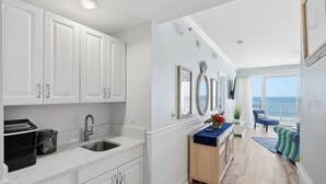 Een moderne keuken met witte kasten, een roestvrijstalen spoelbak en een marmeren aanrecht. Er staat een houten buffetkast met een bloevas. De kamer heeft een groot raam met uitzicht op de oceaan.