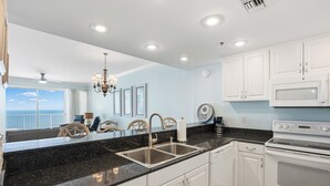 Une cuisine moderne avec des meubles blancs, un plan de travail en granit noir, des éviers en acier inoxydable et des appareils électroménagers blancs, notamment un four à micro-ondes et un four traditionnel.