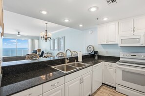 A modern kitchen with white cabinets, a black granite countertop, stainless steel sinks, and white appliances including a microwave and oven. - Princess Condos - All Units Are Gulf Front & Direct Beach Access in PCB! (Panama City Beach)