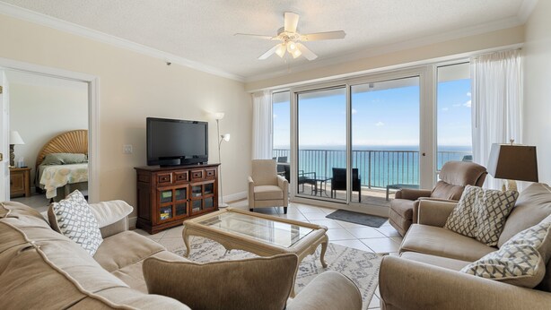 A spacious living room with a large flat-screen TV, comfortable seating, a ceiling fan, and a balcony with a view of the ocean. - Princess Condos - All Units Are Gulf Front & Direct Beach Access in PCB! (Panama City Beach)