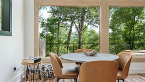 Un espace repas moderne avec une table ronde et quatre chaises, un saladier posé sur la table et une vue sur les arbres à travers de grandes fenêtres.