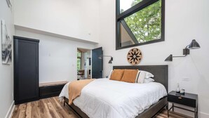 Une chambre moderne avec un grand lit, un plancher en bois, une armoire sombre, un bureau et une fenêtre donnant sur des arbres.