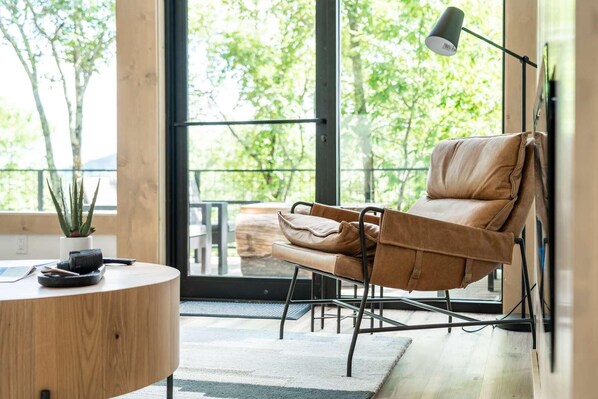 Una habitación moderna con una silla de cuero, una mesa redonda de madera, una planta en maceta y un amplio ventanal con vistas a árboles.