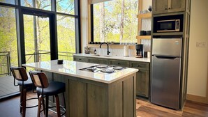 Une cuisine moderne dotée d’un grand îlot central, d’appareils électroménagers en acier inoxydable et offrant une vue sur les arbres grâce à de grandes fenêtres.