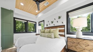 Une chambre à coucher comprenant un lit, des tables de chevet, un ventilateur de plafond, des fenêtres et une vue sur la verdure.