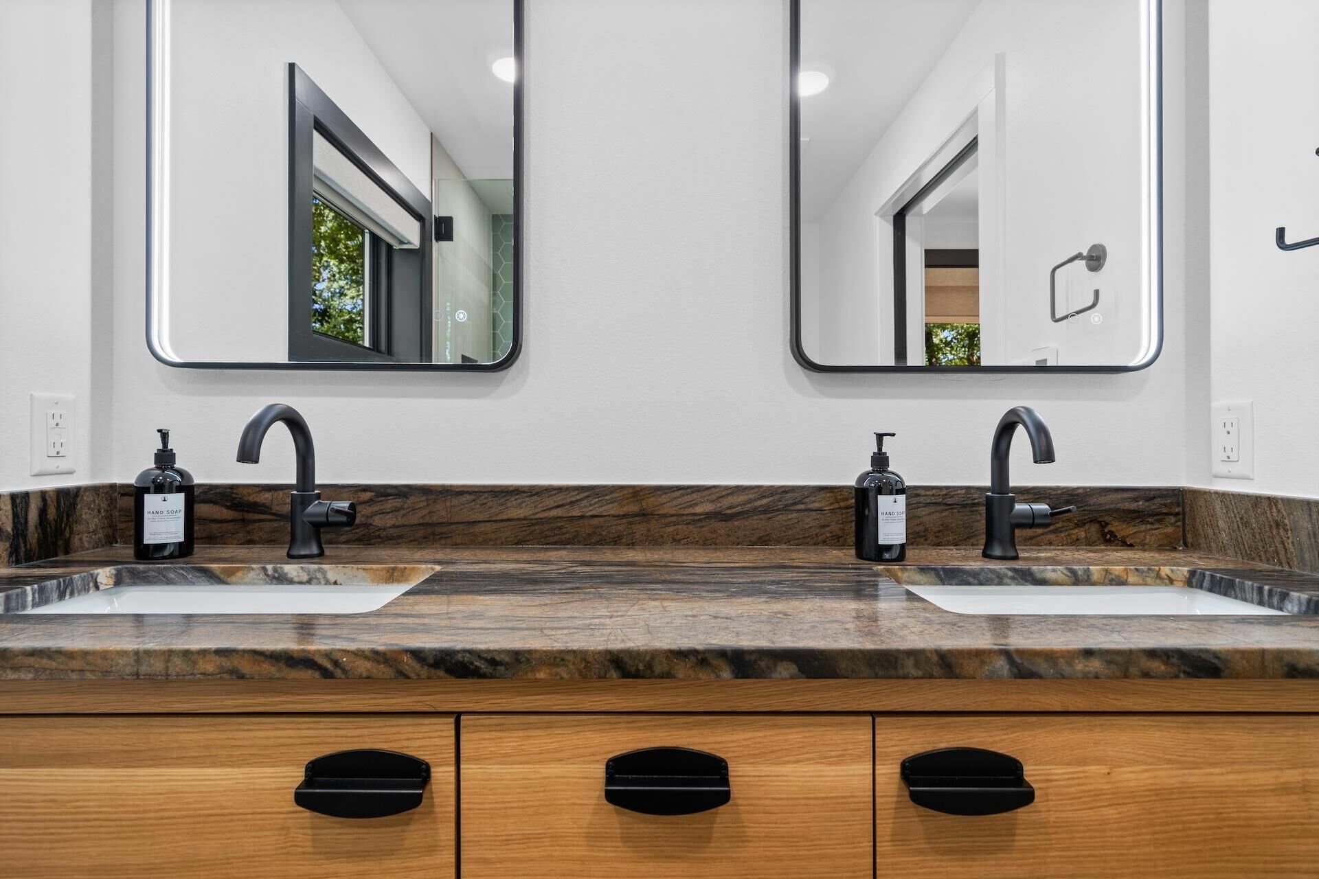 Une salle de bain avec deux lavabos, chacun doté d’un robinet noir et d’un distributeur de savon. On trouve des meubles en bois sous les lavabos.