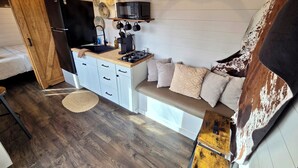 A compact kitchen with a built-in stove, microwave, and sink. There is a wooden cabinet with drawers and a bench with cushions. A rustic horn decorates the wall, and a hanging plant is visible. - The Fox Den - Peaceful Tiny Home Retreat in the Mountains w/ Hot Tub & Fire Pit (Canton)