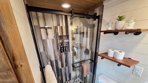A modern bathroom with a glass shower enclosure, a toilet, and wooden shelves with toiletries. - The Fox Den - Peaceful Tiny Home Retreat in the Mountains w/ Hot Tub & Fire Pit (Canton)