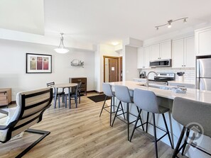 A modern kitchen with white cabinets, a stainless steel refrigerator, a microwave, and a coffee maker. There is a dining area with a wooden table and chairs. - Top-Floor Silver Rock 1BR – Comfort in Every Stay (Fernie)