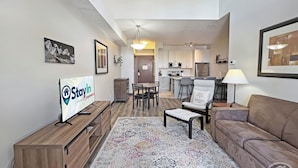 A modern living room with a sofa, a coffee table, and a TV on a wooden stand. There is a dining area with chairs and a table, and a kitchen in the background. - Top-Floor Silver Rock 1BR – Comfort in Every Stay (Fernie)
