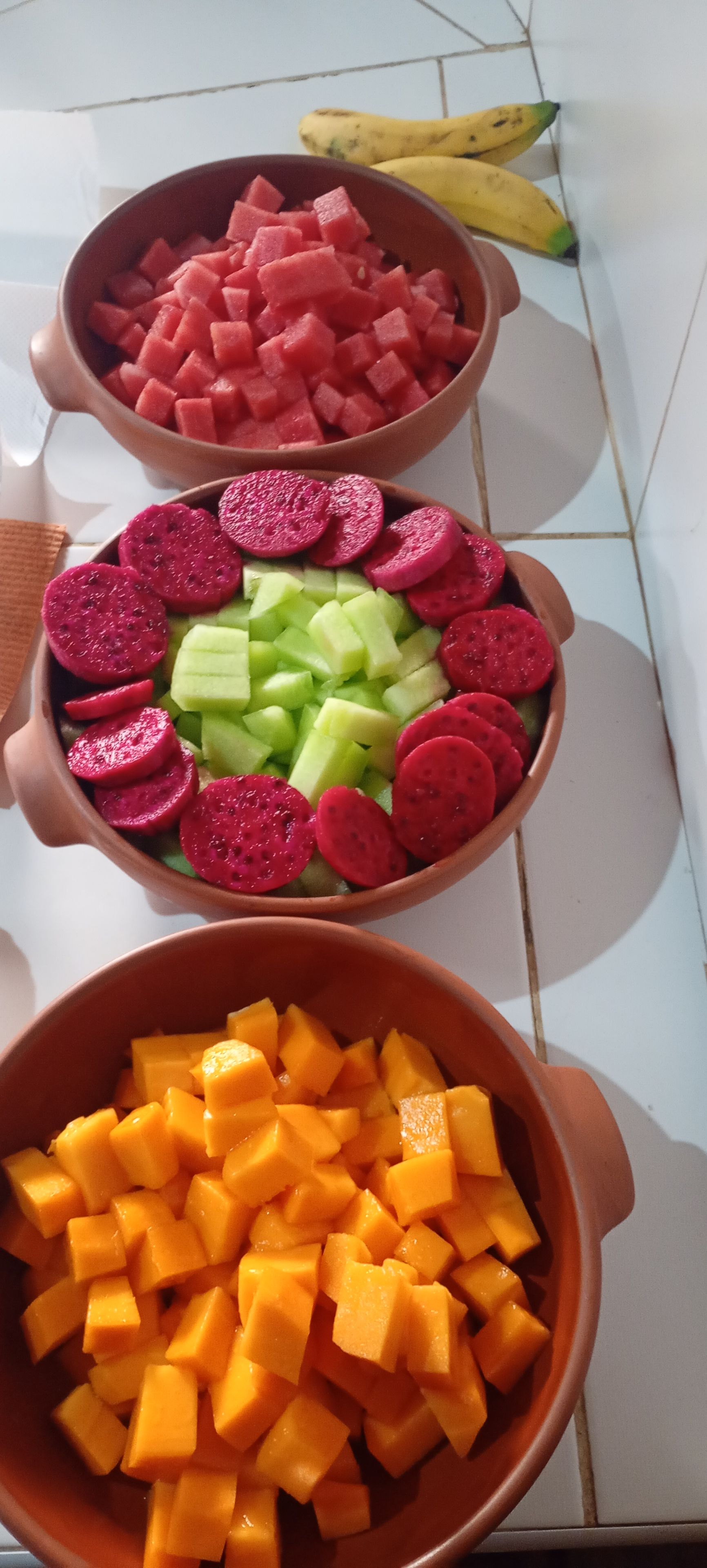 Three ceramic bowls containing diced fruits and vegetables on a tiled floor.