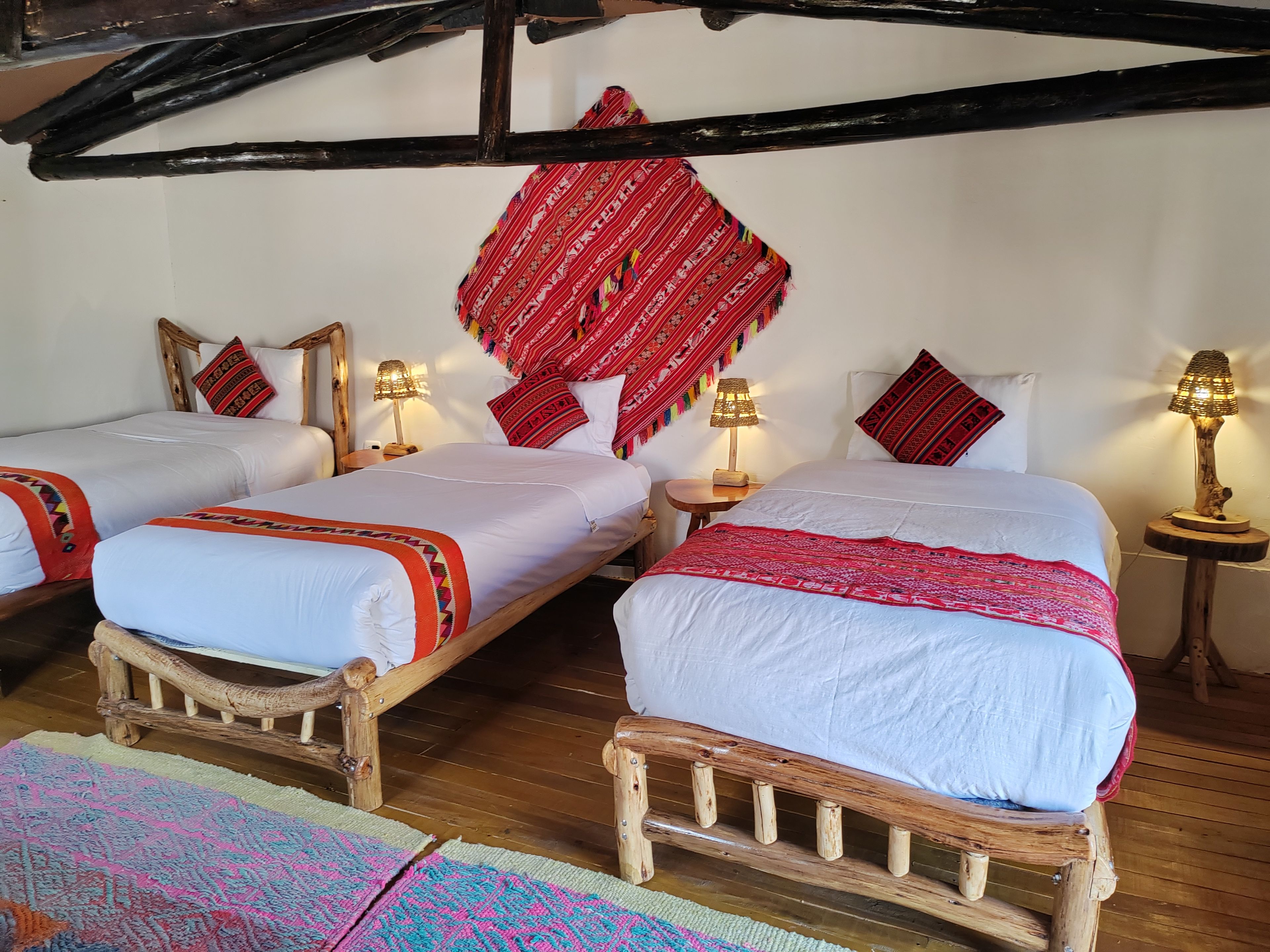 Una habitación con dos camas de madera, cada una con un colchón blanco y colchas rojas estampadas. Hay dos mesitas de noche con lámparas, y un tapiz rojo estampado sobre las camas.